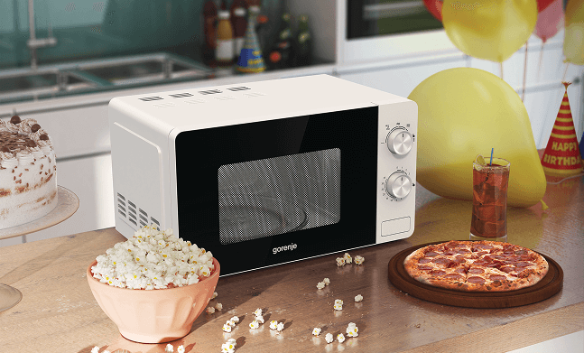 Biała kuchenka mikrofalowa Gorenje na blacie, obok miska popcornu, pizza i szklanka napoju; w tle dekoracje urodzinowe.
