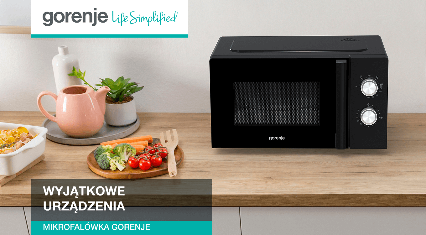 Czarna kuchenka mikrofalowa marki Gorenje stojąca na drewnianym blacie kuchennym. Obok znajdują się warzywa na desce do krojenia oraz ceramiczny dzbanek. W lewym górnym rogu widoczne logo Gorenje z hasłem „Life Simplified”.