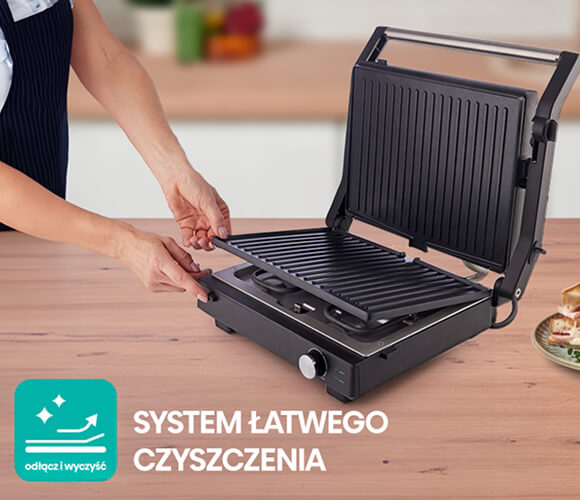 Obrazek przedstawia osobę montującą płytę grilla do czarnego elektrycznego grilla, który znajduje się na drewnianym blacie. Grill jest otwarty, ukazując pionowo ułożone kratki do przygotowywania kanapek. Zarówno kratki, jak i obudowa grilla są w kolorze czarnym. W lewym dolnym rogu obrazu znajduje się turkusowy kwadrat z napisem SYSTEM ŁATWEGO CZYSZCZENIA, co podkreśla funkcję łatwego czyszczenia urządzenia. Po prawej stronie na drewnianym blacie leży talerz z kanapkami, co dodaje apetycznego akcentu do całej sceny.