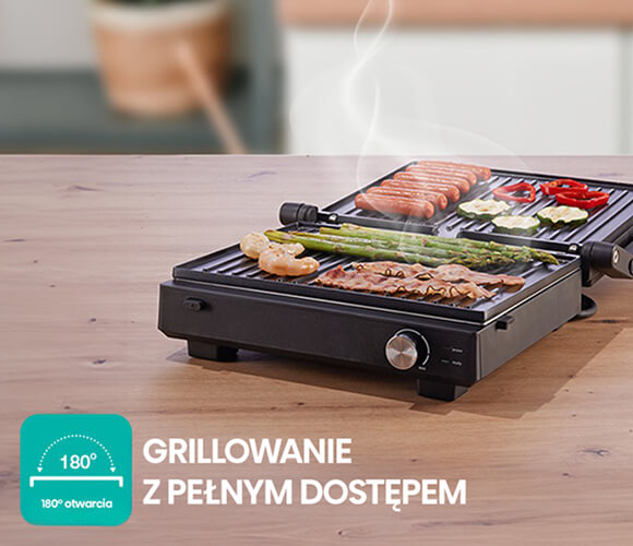 Obrazek przedstawia czarny grill elektryczny umieszczony na drewnianym blacie, który intensywnie gotuje różnorodne potrawy. Na górnej płycie grilla znajdują się kiełbaski oraz pokrojone warzywa, takie jak czerwona papryka i cukinia. Na dolnej płycie grilla leżą zielone szparagi, krewetki oraz plastry boczku. Z potraw unosi się para, co sugeruje, że jedzenie jest gorące i świeżo przygotowane. W lewym dolnym rogu znajduje się zielony kwadrat z zaokrąglonymi rogami, w którym widnieje stylizowany symbol półokręgu z napisem 180° oraz tekst 180 otwarcia poniżej. Obok kwadratu znajduje się napis GRILLOWANIE Z PEŁNYM DOSTĘPEM, co podkreśla funkcjonalność grilla.