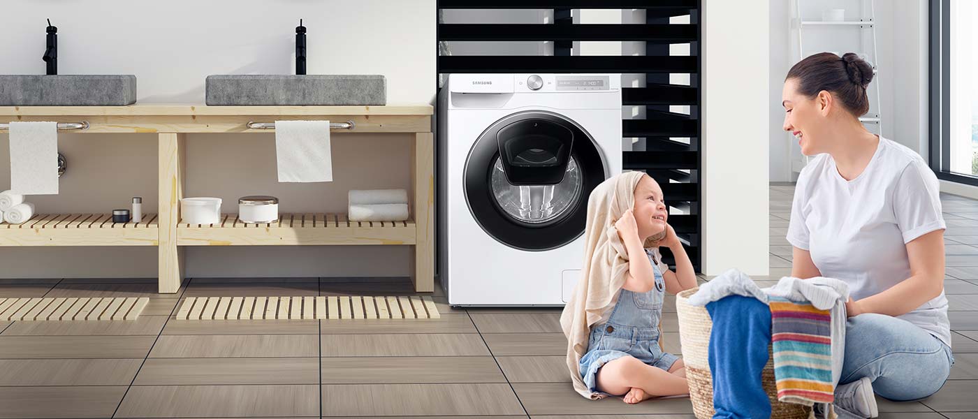 Mama i córka bawią się na podłodze łazienki koszem z ubraniami, za nimi pralka Samsung i drewniane meble z kamiennymi umywalkami