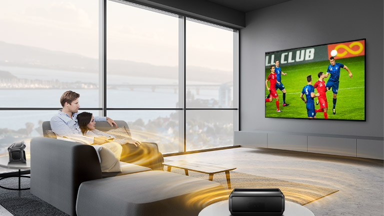 Mężczyzna i kobiety oglądają sport w telewizji w salonie z podłączonymi tylnymi głośnikami Bluetooth