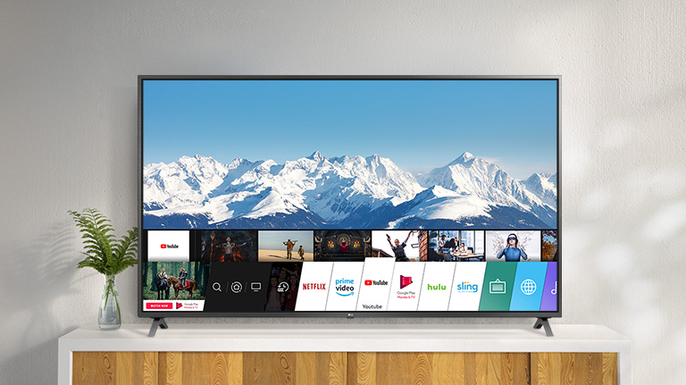 Telewizor stojący na białej podstawie na tle białej ściany. Ekran telewizora z wyświetlonym ekranem głównym systemu webOS.