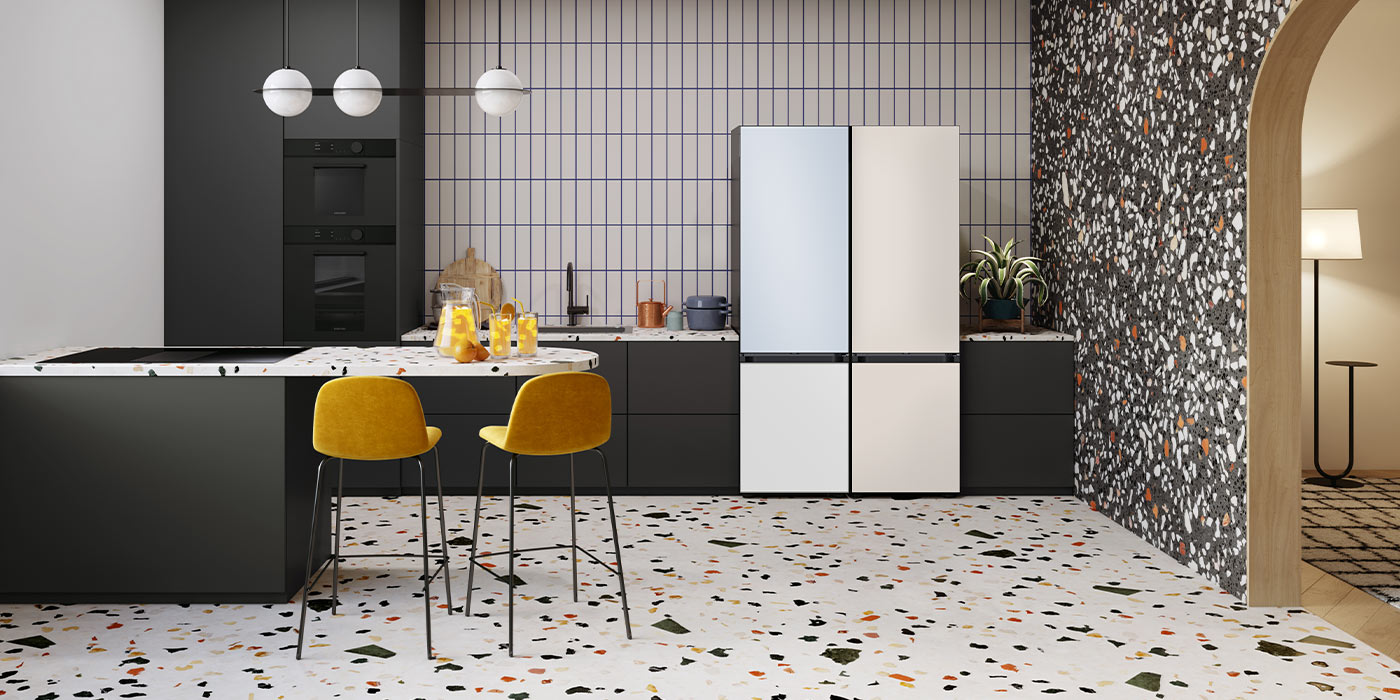 Para lodówek Samsung BESPOKE, stojących w pomieszczeniu kuchennym z ciemnymi meblami. Jedna z lodówek posiada panel drzwiowy w kolorze kremowego błękitu, a druga alabastrowej bieli