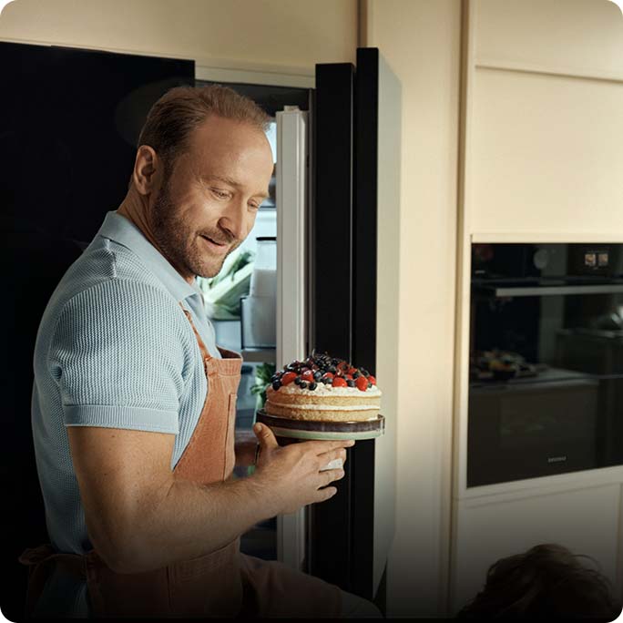 Uśmiechnięty Borys Szyc w fartuchu kuchennym wyjmuje z lodówki mały tort z owocami.