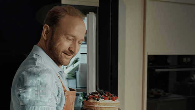 Uśmiechnięty Borys Szyc w fartuchu kuchennym wyjmuje z lodówki mały tort z owocami.