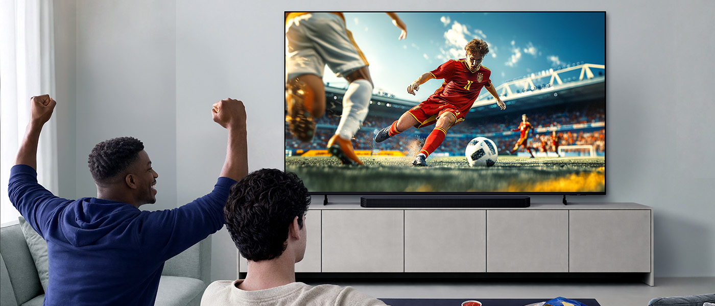 Dwóch mężczyzn siedzi w jasnym pokoju i ogląda na telewizorze mecz piłkarski, gdy jeden z nich podnosi się z radości. 